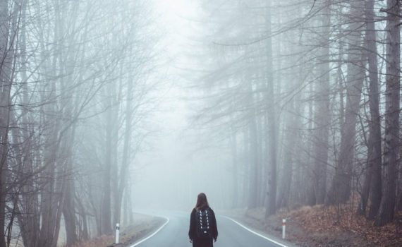 Person walks alone on foggy forest road, surrounded by tall trees, creating a serene and mysterious atmosphere.