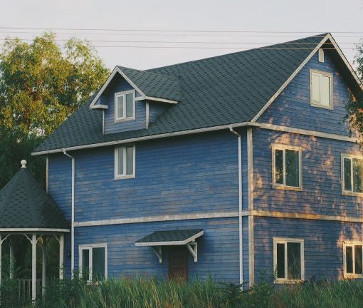 Blue wooden house with a gazebo surrounded by trees and greenery under a clear sky.
