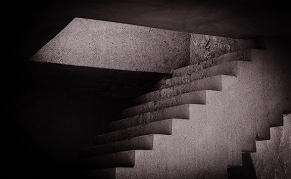 Monochrome abstract image of a staircase with dramatic lighting from an opening above, creating geometric shadow patterns.