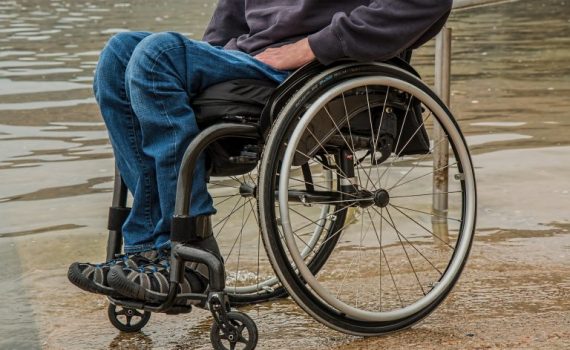 Person in a wheelchair by water, wearing casual clothes and sneakers, on a concrete path.