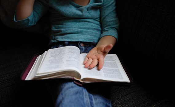 Child reading an open book on lap, wearing casual teal shirt and jeans, sitting on a couch, hand gesturing thoughtfully.