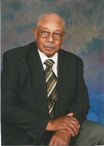 Elderly man in a suit with a tie, sitting against a blue and gray backdrop, looking towards the camera.