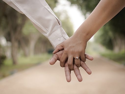 Close-up of a couple holding hands outdoors, highlighting a diamond engagement ring.