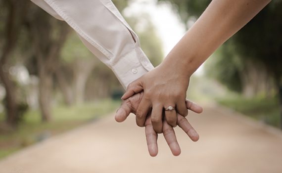 Close-up of a couple holding hands outdoors, highlighting a diamond engagement ring.