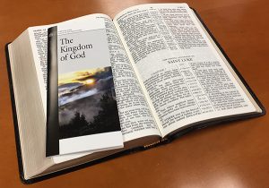 Open Bible with The Kingdom of God pamphlet on a wooden table, emphasizing spiritual study and reflection.
