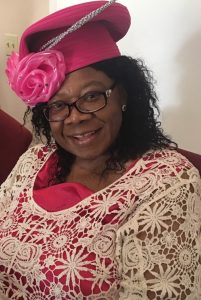 Elegant woman in pink hat with rose detail, wearing crochet lace top, sitting and smiling warmly indoors.