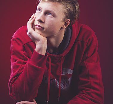 Young person in red hoodie rests chin on hand, gazing upwards thoughtfully against a red background.