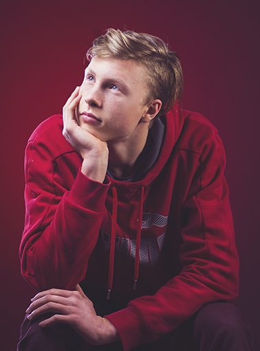 Young person in red hoodie rests chin on hand, gazing upwards thoughtfully against a red background.