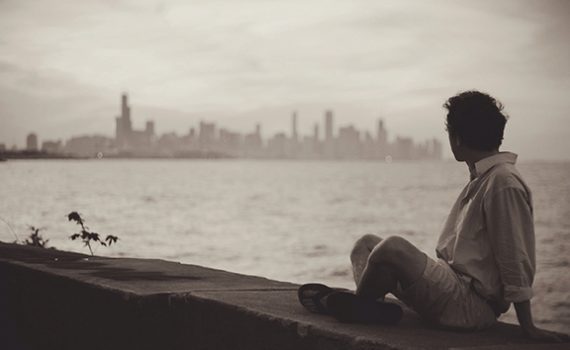 Person sitting by waterfront, gazing at distant city skyline in sepia tones. Urban landscape and atmosphere.