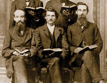 Vintage photo of five men in suits holding open books, seated and standing, sepia tone, 19th-century style.