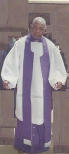 Elderly man in white and purple church robes, standing indoors, facing forward.