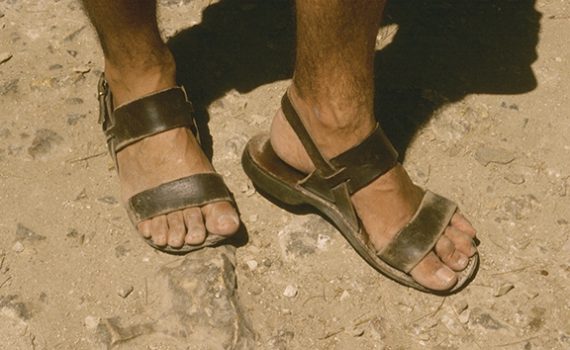 Worn leather sandals on feet, standing on a rocky, sunlit path.