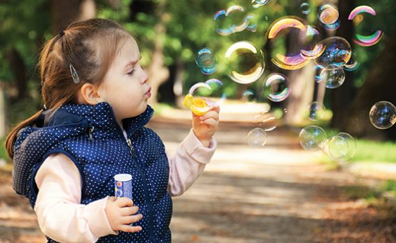 Child blowing soap bubbles outdoors in a park, wearing a blue vest, with trees in the background.