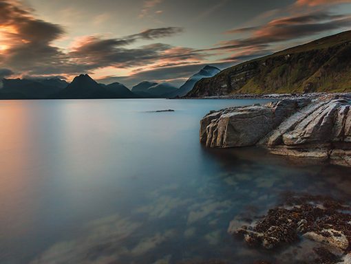 Tranquil sunset over a serene lake with rocky shoreline and distant mountains, under a colorful sky.