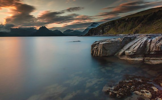 Tranquil sunset over a serene lake with rocky shoreline and distant mountains, under a colorful sky.