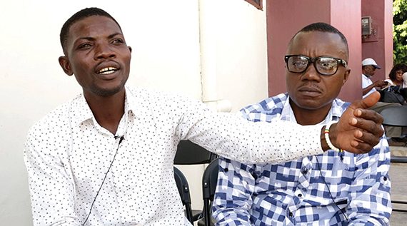 Two men in conversation outdoors; one gestures, wearing a white shirt, the other listens in glasses and a checked shirt.