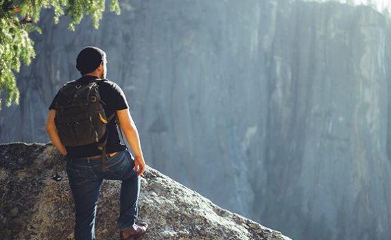 Man with backpack standing on rocky cliff, overlooking a vast natural landscape. Adventure and exploration concept.