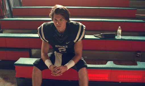 Football player in uniform sits on bleachers, focused and contemplative. Sports equipment visible. Athletic scene.