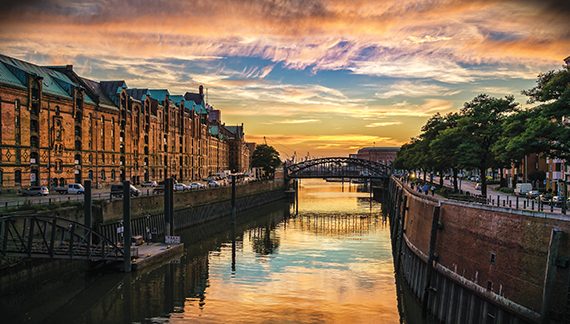 Sunset over a historic canal with old brick buildings and a bridge reflecting in the water.