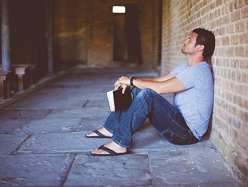Man sitting against brick wall holding book, appearing contemplative in outdoor corridor setting.