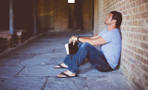 Man sitting against brick wall holding book, appearing contemplative in outdoor corridor setting.