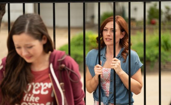 Woman standing behind black fence bars, watching a person in a red shirt walk away in a garden setting.
