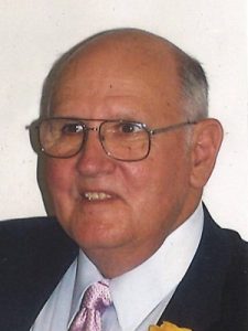 Elderly man in glasses and suit with a pink tie, smiling slightly, against a plain background.