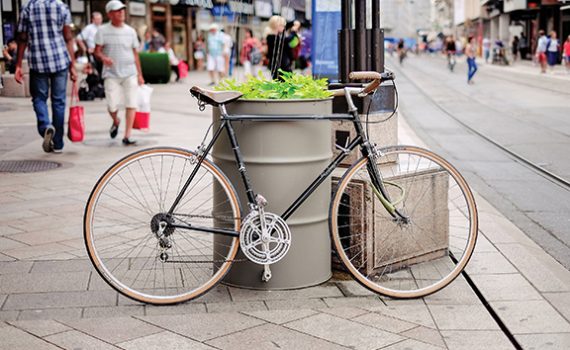Bicycle parked by a large planter on a busy city street with people walking and shops in the background.