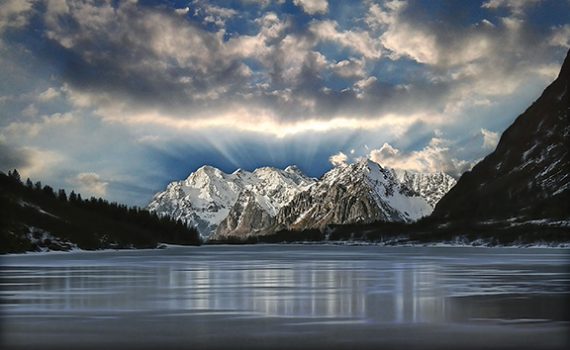 Snow-capped mountains under a dramatic sky with clouds, reflected in a calm, icy lake at sunrise.