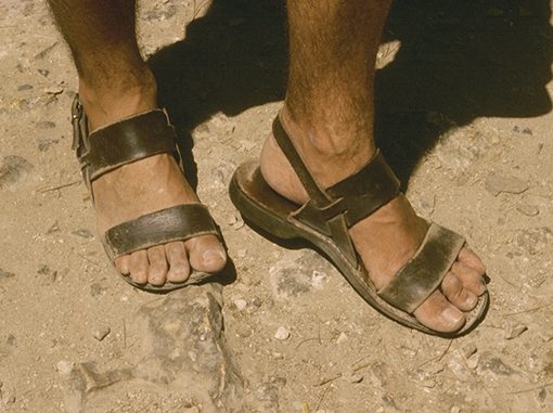 Worn leather sandals on a dusty ground, showing feet and sunlit legs, evoking a rustic outdoor setting.