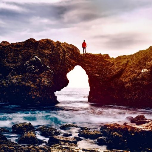 Person standing on a natural rock arch over ocean waves, with a cloudy sky overhead, creating a scenic coastal view.