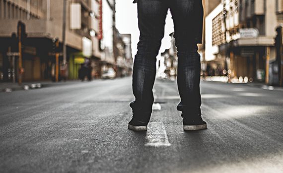 Person in jeans and sneakers standing on an empty city street, viewed from behind, with buildings lining the road.