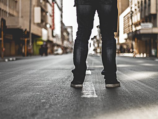 Person in jeans standing on an empty urban street, back view, with buildings lining the road.