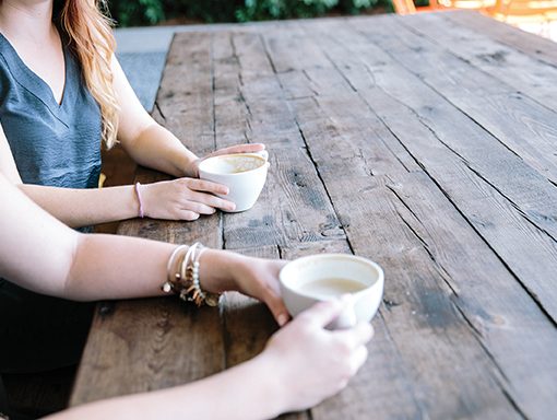Two people holding coffee cups at a rustic wooden table, engaged in a casual conversation outdoors.