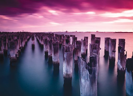 Sunset over old wooden pier posts in tranquil water, creating a serene and colorful seascape scene.