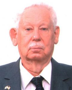 Elderly man in a suit with white hair and mustache, posing against a plain background.