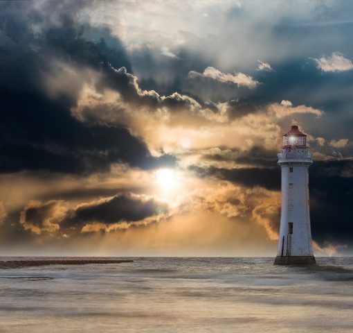 Lighthouse by the sea at sunset with dramatic clouds and sunlight breaking through, creating a serene coastal scene.