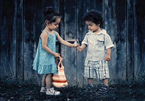 Two children standing together, one holding a basket, sharing a moment outdoors in front of a wooden fence.
