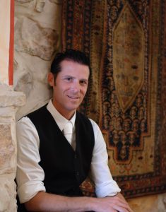 Man in black vest and white shirt sitting by a stone wall with an ornate rug in the background.