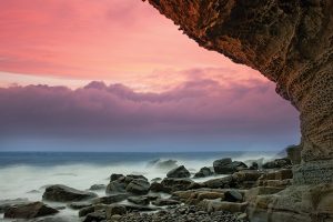 Sunset over rocky shore with dramatic pink clouds and ocean waves under a cliff.