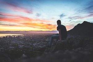 Person sitting on rock, overlooking cityscape at sunset with colorful sky and mountains in the distance.