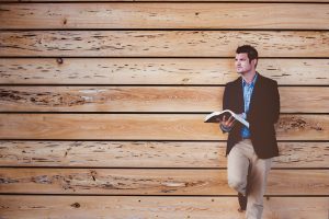 Man reading a book while leaning on a wooden wall, wearing a blazer and jeans, with a contemplative expression.