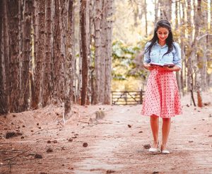 Woman reading a book while walking on a forest path, surrounded by tall trees in autumn.