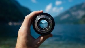 Hand holding camera lens with scenic mountain and lake view, focusing on nature through photography.