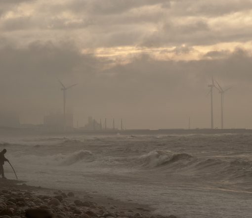 Person walking on misty beach near sea with wind turbines in the background.