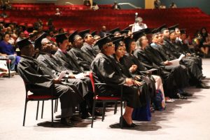 Graduates in caps and gowns seated at a commencement ceremony in an auditorium, ready to receive diplomas.