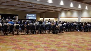 Large conference audience seated in a spacious hall with presentation screen, professional gathering.