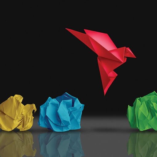 Red origami bird flying above colorful crumpled paper balls on a reflective surface.