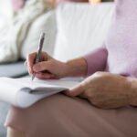 Person writing in a notebook on their lap while sitting on a couch, wearing a pink sweater.